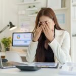 A woman looking mentally tired at her desk with papers, a laptop, and a phone