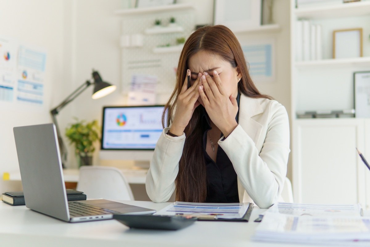 A woman looking mentally tired at her desk with papers, a laptop, and a phone